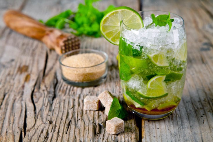 A mojito sitting on a table surrounded by sugar, mint, and lime