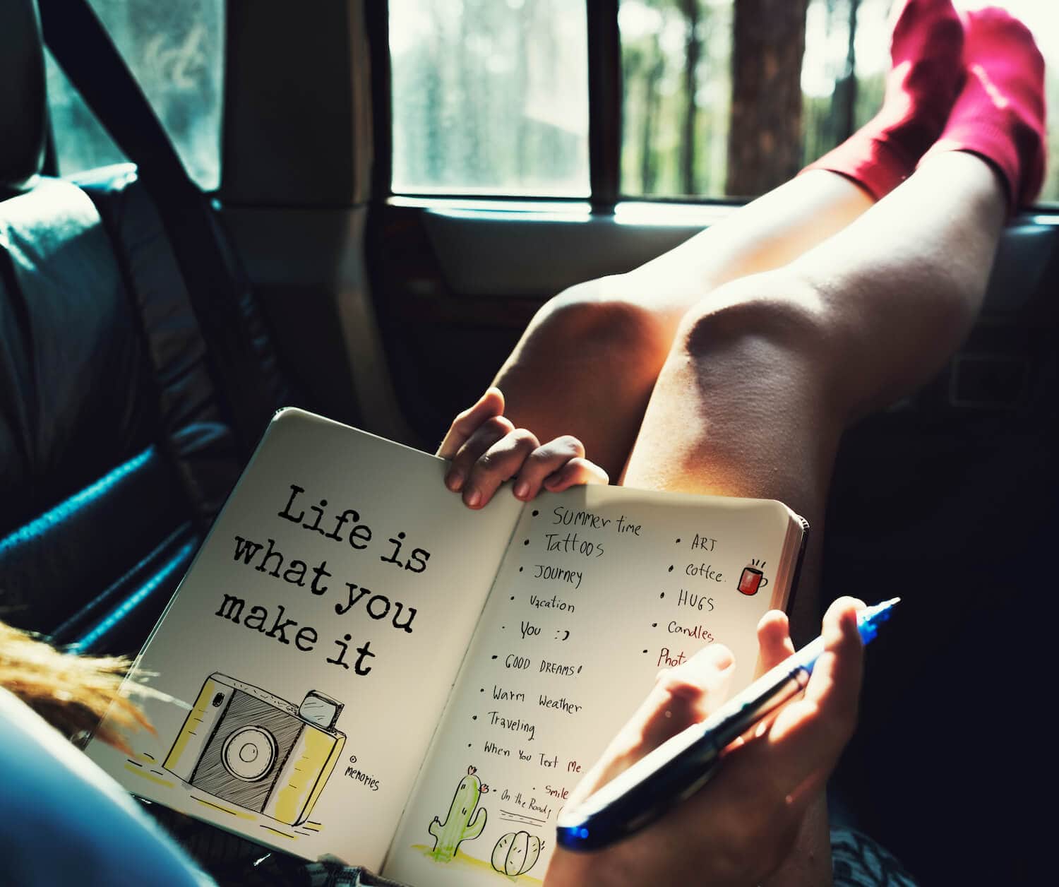 A woman with her legs up on the window of a car while she writes in a travel journal.