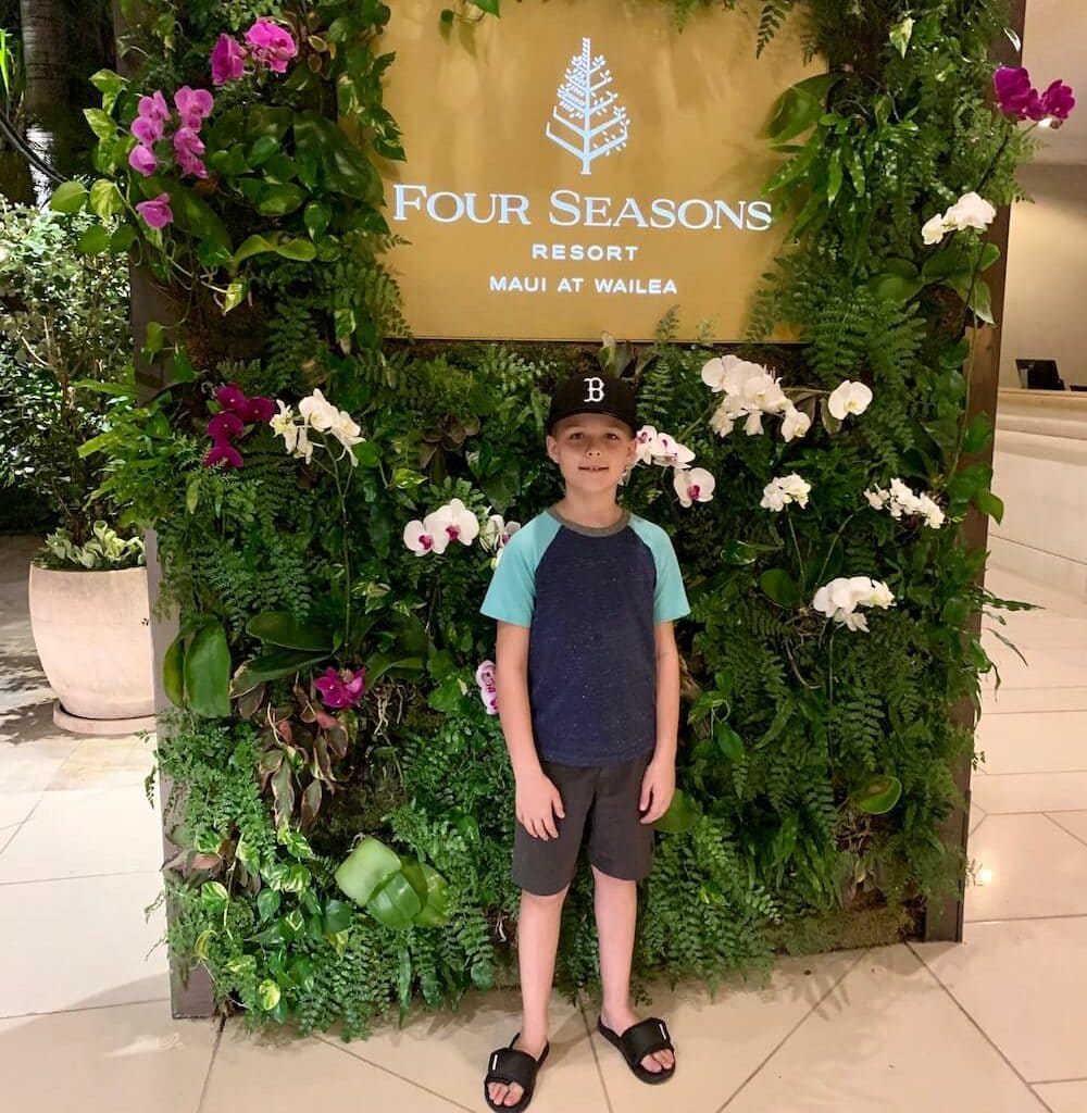 A boy in front of a lush green sign of the Four Season Resort in Maui. 